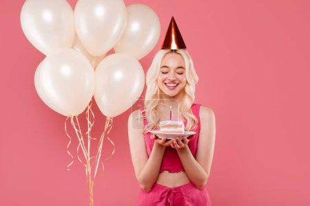 Photo for Pretty lady in hat holding piece of cake and making wish for birthday, posing near inflatable balloons isolated on pink background, studio. Emotions from celebrating holiday - Royalty Free Image