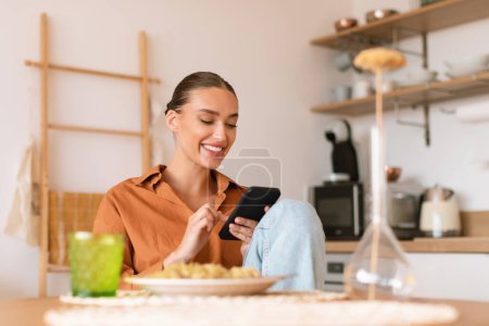 Photo for Excited lady using smartphone in kitchen while having dinner, sitting at table and texting on phone, browsing social networks or shopping online, free space - Royalty Free Image