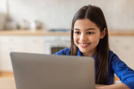 Foto de Estudiando en línea. Retrato de cerca de la alegre chica árabe sentada en el escritorio con computadora portátil en su habitación, usando PC portátil, chat y navegación web, trabajando en el proyecto educativo en casa - Imagen libre de derechos