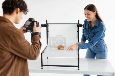 Photo for Team of male photographer and his female assistant doing content photoshoot for fast food restaurant, taking photos of burger on white platform, woman helping while working in team in photostudio - Royalty Free Image