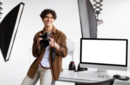 Téléchargez les photos : Photographe européen joyeux homme avec caméra dslr assis près de l'écran de l'ordinateur avec écran vierge de travail en photostudio, modèle. Homme avec caméra posant et souriant à la caméra - en image libre de droit