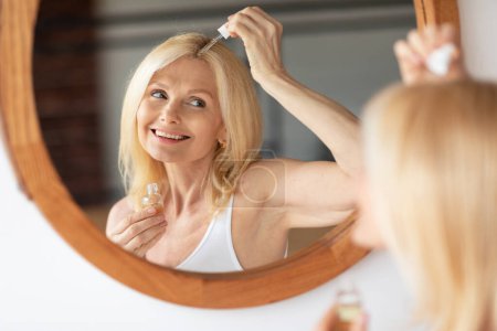 Photo for Happy mature woman applying serum or natural oil on her scalp, looking at her reflection in mirror and smiling. Organic cosmetics for haircare, damaged dry hair treatment, daily beauty routine - Royalty Free Image