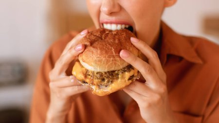 Téléchargez les photos : Femme caucasienne affamée aiment manger hamburger savoureux, mordre maison préparé ou livré repas malbouffe, gros plan, cultivé, panorama. Concept de malbouffe - en image libre de droit