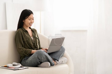 Photo for Smiling beautiful millennial chinese woman wearing casual outfit sitting on couch with computer laptop on his lap, typing on pc keyboard, home interior, copy space. Chatbot, AI in daily life - Royalty Free Image
