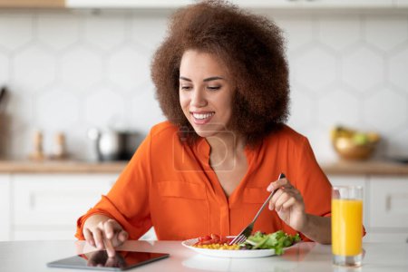 Foto de Mujer negra alegre con tableta digital Noticias de lectura durante el desayuno en la cocina, Smiling African American Female Sitting at Table, Disfrutando de la comida sabrosa y navegar por Internet, Espacio libre - Imagen libre de derechos