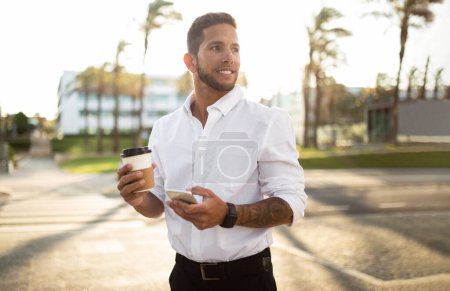 Téléchargez les photos : Heureux jeune homme entrepreneur tenant café à emporter et en utilisant un téléphone portable tout en marchant à l'extérieur pendant la pause et passer du temps à l'extérieur, copier l'espace - en image libre de droit