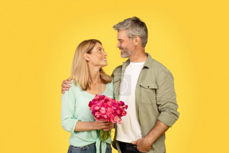Photo for Loving european spouses hugging and smiling at each other, woman holding bouquet of flowers, enjoying romantic, posing on yellow background. Congratulations of holiday, Valentine day and anniversary - Royalty Free Image