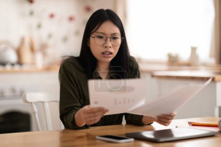 Photo for Distressed overwhelmed pretty young asian woman in casual wearing eyeglasses sitting at kicthen desk with gadgets laptop and phone on, checking papers or correspondence, have difficulties at work - Royalty Free Image