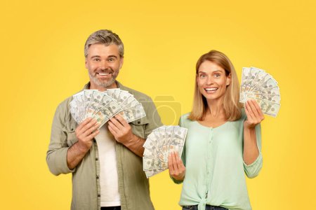 Photo for Family savings concept. Happy middle aged couple standing isolated over yellow studio background, showing money dollar banknotes, looking at camera and smiling - Royalty Free Image