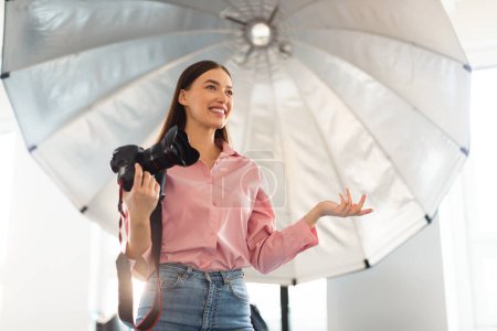 Photo for Professional photographer woman with dslr camera standing in front of reflective umbrella, working in modern photostudio and explaining something, free space - Royalty Free Image