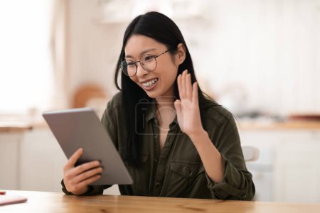 Photo for Telecommunication concept. Cheerful happy young chinese woman wearing casual outfit and eyeglasses using digital tablet at kitchen, waving at pad screen, have video chat with friends, copy space - Royalty Free Image
