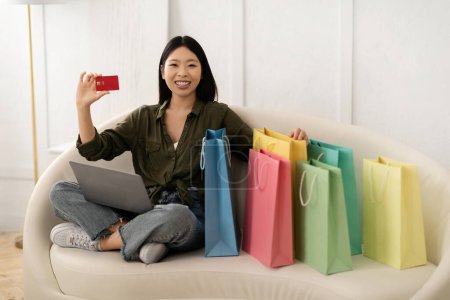 Photo for Happy beautiful young asian woman in casual outfit sitting on couch among colorful shopping bags, using bank card and laptop pc computer, customer shopping online from home. E-commerce, retail - Royalty Free Image