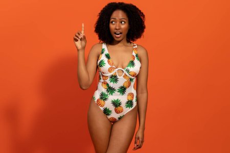 Photo for Nice ideas for summer vacation concept. Inspired happy pretty youg black woman in swimsuit with pineapples raising finger up, isolated on orange studio background, copy space for offer advertisement - Royalty Free Image
