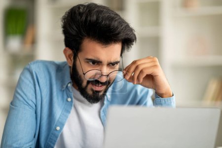 Photo for Indian Freelancer Guy Taking Off Glasses And Looking At Laptop Screen In Shock, Confused Eastern Man Reading Unpleasant Email Or Checking Business News While Working On Computer At Home Office - Royalty Free Image