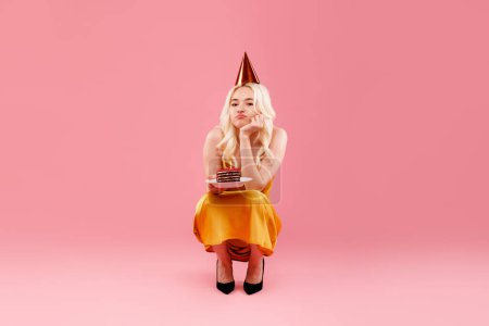Foto de Triste joven con sombrero de fiesta sosteniendo pastel de cumpleaños, mirando a la cámara con una profunda mueca molesta, sintiéndose sola en su cumpleaños, sentada sobre un fondo rosa - Imagen libre de derechos
