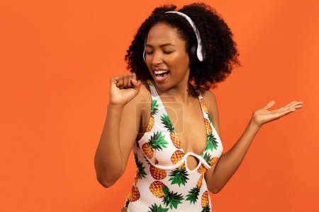 Téléchargez les photos : Joyeux jolie jeune femme noire bouclée dans des maillots de bain d'été élégants en utilisant des écouteurs sans fil et en dansant en bougeant son corps sur fond orange, en écoutant de la musique et en chantant pendant les vacances, en copiant l'espace - en image libre de droit