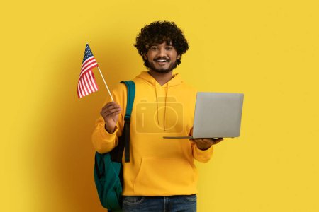 Foto de Feliz joven oriental con capucha amarilla llevando mochila, estudiante hindú sosteniendo la computadora portátil y la bandera de los Estados Unidos, aislado en el fondo amarillo. Educación en el extranjero, estudio en América concepto - Imagen libre de derechos