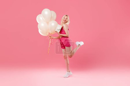 Téléchargez les photos : Joyeux anniversaire de la jeune femme, tenant un tas de ballons et boîte cadeau avec ruban cadeau arc, posant isolé sur fond de studio rose, plan complet - en image libre de droit