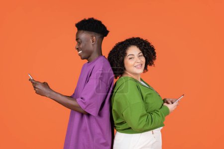 Photo for Cheerful handsome young black guy and hispanic lady plus size standing back to back, using phones, websurfing, scrolling on social media, isolated on orange background - Royalty Free Image