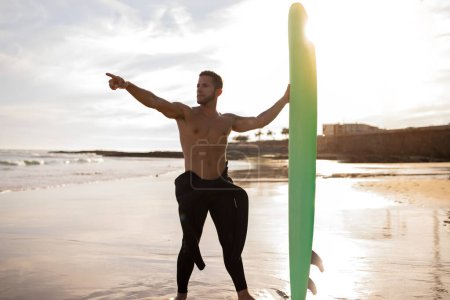 Photo for Water Sports Concept. Handsome Young Guy Surfing On The Beach At Sunset, Muscular Surfer Man Standing With Surfboard And Pointing Aside, Millennial Male In Wetsuit Ready To Ride Waves, Copy Space - Royalty Free Image