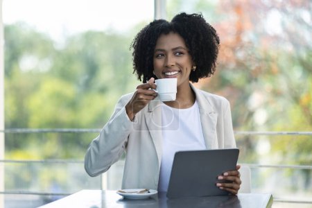 Foto de Mujer negra bastante joven que usa ropa informal inteligente empresaria sentada en la cafetería, usando tableta digital, bebiendo café, trabajando en un proyecto de negocios, mirando a un lado y sonriendo, espacio para copiar - Imagen libre de derechos