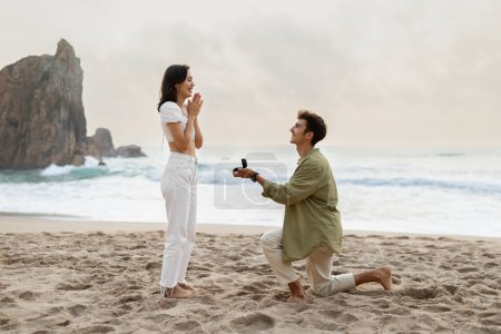 Foto de Propuesta romántica en la orilla del mar. Joven amante con anillo de compromiso haciendo propuesta a la mujer feliz en la playa en la costa, vista lateral - Imagen libre de derechos