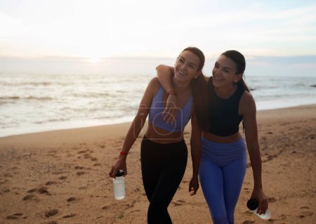Téléchargez les photos : Bonnes jeunes femmes européennes jumelles en vêtements de sport câlins, repos après l'entraînement avec des bouteilles d'eau sur la plage de la mer, en plein air. Entraînement ensemble, mode de vie actif, jogging le matin au lever du soleil - en image libre de droit