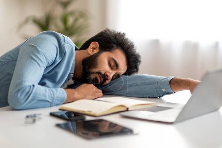 Foto de Concepto de Procrastinación. Hombre indio joven freelancer durmiendo en el escritorio con el ordenador portátil, cansado Millennial Oriental Guy siesta en la mesa, tener descanso en el trabajo, sentirse agotado después de trabajar en la computadora - Imagen libre de derechos