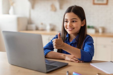 Téléchargez les photos : En ligne Homeschooling. Portrait de sourire Preteen Middle Eastern Girl Étudier avec ordinateur portable, Gesturing Thumbs Up assis près du bureau à la maison. Enfant montrant comme geste au PC Approuver la leçon - en image libre de droit