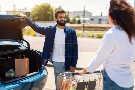 Photo for Happy arab couple with shopping cart full of fresh food, packing products into the car on the outdoor parking, woman standing near shopping cart - Royalty Free Image