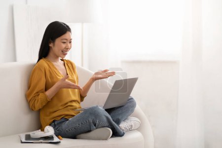 Photo for Positive smiling young asian woman in casual outfit project manager or accountant working from home, sitting on couch, using laptop, have online meeting with colleagues, copy space - Royalty Free Image