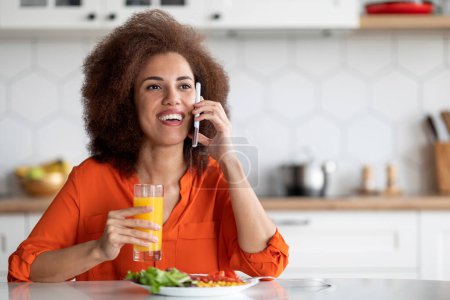 Foto de Alegre mujer negra hablando en el teléfono móvil y comiendo el desayuno en la cocina, feliz joven afroamericana sentada en la mesa y disfrutando de una agradable conversación en el teléfono celular, espacio de copia - Imagen libre de derechos