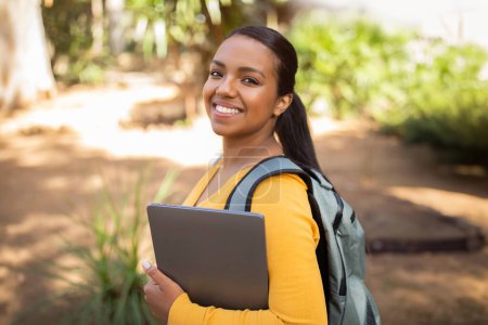 Téléchargez les photos : Portrait d'une étudiante hispanique heureuse avec ordinateur portable et sac à dos debout à l'extérieur dans le parc, souriant à la caméra. Jeune femme tenant ordinateur portable posant à l'extérieur après les cours universitaires - en image libre de droit