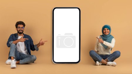 Téléchargez les photos : Heureux couple musulman assis et pointant vers grand smartphone avec écran blanc pour la maquette, recommander une nouvelle application, homme arabe joyeux et femme en hijab posant avec téléphone portable sur fond beige - en image libre de droit