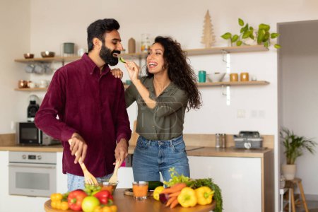Foto de Jóvenes cónyuges árabes felices preparando deliciosa comida juntos, amando a la mujer que alimenta a su marido mientras cocina en casa, divirtiéndose, interior de la cocina, espacio libre - Imagen libre de derechos