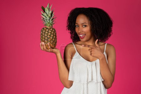 Photo for Happy young black curly lady pointing finger at pineapple, isolated on pink background, studio. Fun, summer fruit recommendation, emotions and diet, lifestyle, ad and offer - Royalty Free Image
