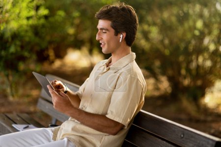 Téléchargez les photos : Happy relaxé beau gars en tenue décontractée assis sur un banc à l'extérieur dans la rue, en utilisant tablette numérique et écouteurs, espace de copie. Homme millénaire reposant dans un parc public, regardant du contenu vidéo - en image libre de droit