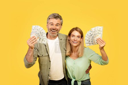 Téléchargez les photos : Joyeux couple européen d'âge moyen tenant les fans d'argent comptant, souriant à la caméra sur fond de studio jaune. Conjoints caucasiens célébrant grande loterie ou casino gagner - en image libre de droit