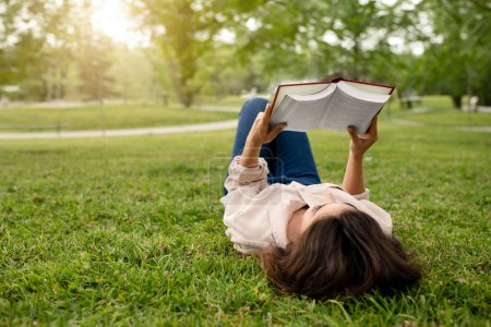 Photo for Student Young Lady Reading Book Lying On Green Grass Outdoor In Campus Park, Relaxing Enjoying Sunny Summer Weekend. Reader Woman Relaxing And Learning Outside. Empty Space. Knowledge And Education - Royalty Free Image