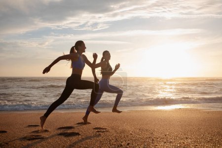 Photo for Happy young slim european women twins in sportswear run at morning, freeze in air at sea beach, outdoor, full length. Cardio workout together, active lifestyle at summer, body care and weight loss - Royalty Free Image
