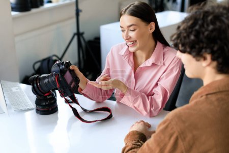 Téléchargez les photos : Photographe femme heureuse montrant des photos après le tournage, discuter du travail avec l'homme en studio, parler après la séance photo et choisir les meilleures photos, assis sur le lieu de travail - en image libre de droit