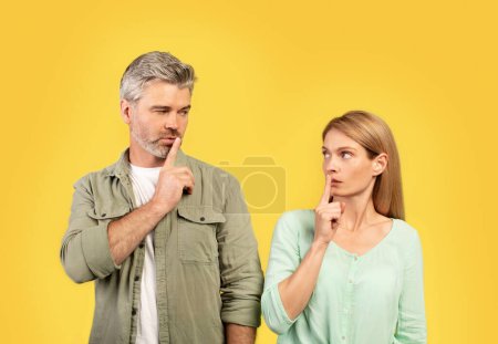 Téléchargez les photos : Chut, tais-toi. Couple européen d'âge moyen gesticulant signe de silence, debout sur fond de studio jaune, se regardant et tenant le doigt sur les lèvres - en image libre de droit