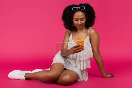 Photo for Happy young black curly lady typing on smartphone, chatting in social networks, sit on floor, isolated on pink background, studio. Communication app, lifestyle, ad and offer - Royalty Free Image
