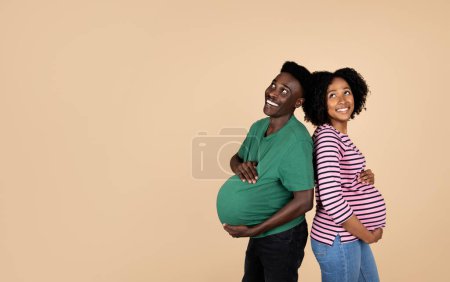 Téléchargez les photos : Heureux jeune mari afro-américain et femme enceinte avec gros ventre dans occasionnel s'amuser ensemble, profiter d'attendre bébé isolé sur fond de studio beige. Amour, famille et relation - en image libre de droit