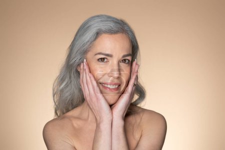 Téléchargez les photos : Portrait d'une femme sensuelle aux cheveux gris âgée touchant son visage et souriant à la caméra, posant sur fond de studio beige. Dame âgée à la peau douce bénéficiant d'un traitement spa - en image libre de droit