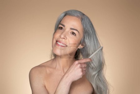 Foto de Mujer mayor de pelo gris sonriente cepillándose el pelo largo y hermoso después de la ducha, posando sobre fondo beige del estudio, espacio para copiar. Cuidado del cabello, concepto de cabello sano y hermoso - Imagen libre de derechos
