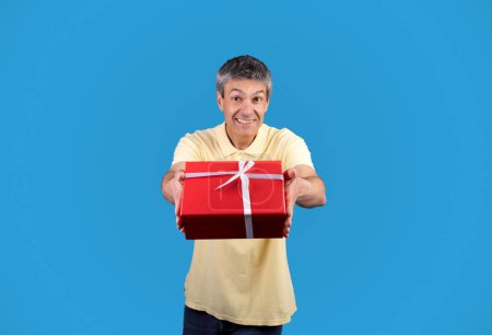 Téléchargez les photos : Présent pour vous. joyeux mature homme donnant enveloppé boîte cadeau souriant et étirant les mains à la caméra, vous félicitant pour anniversaire vacances debout sur fond bleu, Studio Shot - en image libre de droit