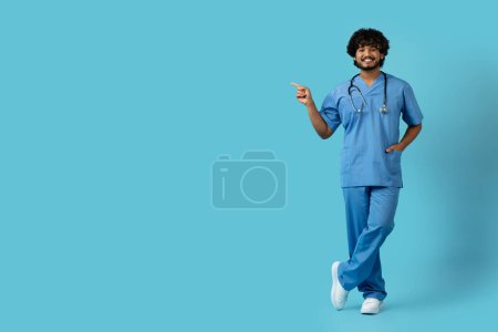 Photo for Doctors, medical staff, healthcare workers. Friendly cheerful handsome curly young eastern man in medical workwear with stethoscope doctor pointing at copy space for ad or text, blue background - Royalty Free Image