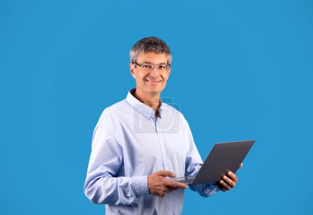 Photo for Online Offer. Happy Mature Man Holding Laptop Computer, Posing Wearing Shirt And Eyeglasses, Advertising Website Standing On Blue Studio Background. Gray Businessman Browsing Internet Via PC - Royalty Free Image