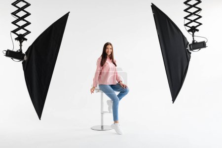 Téléchargez les photos : Joyeux modèle européen dame en tenue décontractée assis sur la chaise et posant sur fond blanc en photostudio, souriant à la caméra, ayant photoshoot et faire du contenu pour la boutique, pleine longueur - en image libre de droit